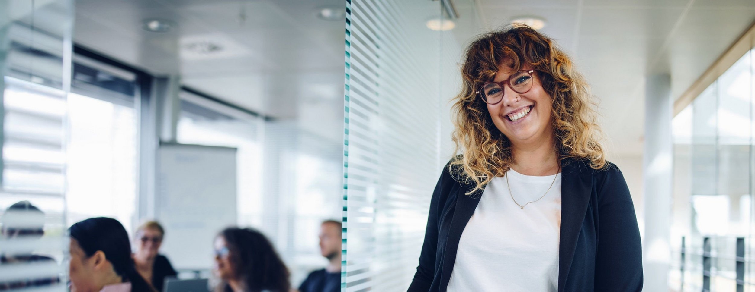 Eine Junge Frau steht neben der offen Tür eines Besprechungsraums mit einigen Personen darin und lächelt in die Kamera.